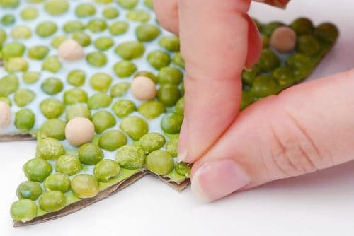 Close-up of fingers adding green peas to glue on cardboard tree shape